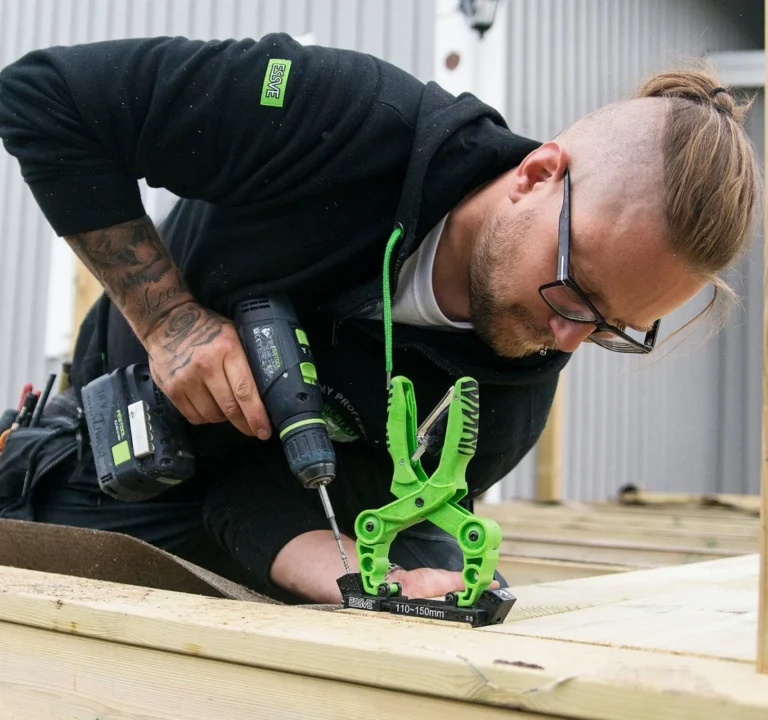 En snickare som använder en skruvdragare och en grön tving under ett husbygge.
